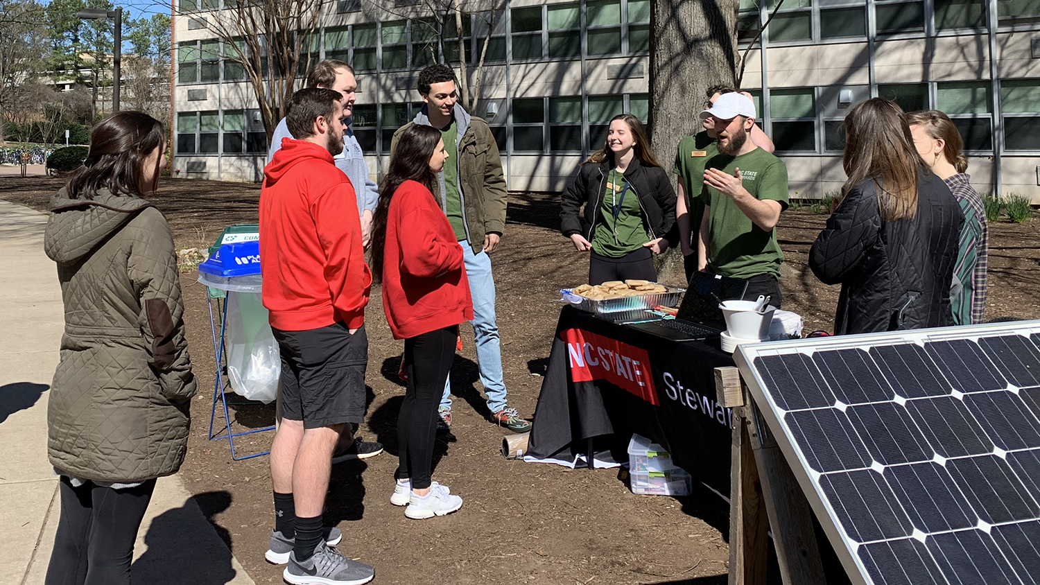 NC State Stewards, wearing matching green T-shirts, speak to a small group of fellow students outside next to a solar panel - Finding Your Wolfpack: Floursih With Sustainability - Forest Biomaterials NC State University