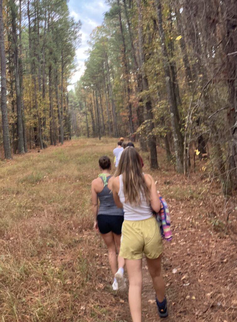 The image shows a group of people walking along a forest trail. Tall trees with autumn-colored leaves line both sides, and the ground is covered in grass and fallen leaves. The group is moving away from the camera.