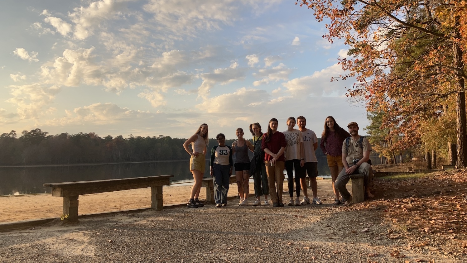 A group of students stand together by a lake during autumn, with warm sunset light and colorful fall trees in the background. - FER GSA Group Camping Trip - Forestry and Environmental Resources NC State University
