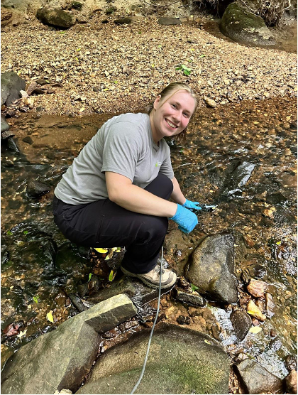 The image shows a woman kneeling beside a shallow rocky stream in an outdoor, wooded area. She is smiling at the camera and wearing a gray T-shirt, dark pants, sturdy outdoor shoes, and blue protective gloves. She appears to be working with a tool or instrument in the water, possibly collecting a sample or taking measurements. The setting is natural, with rocks, flowing water, and trees surrounding her, suggesting fieldwork or an environmental research activity.