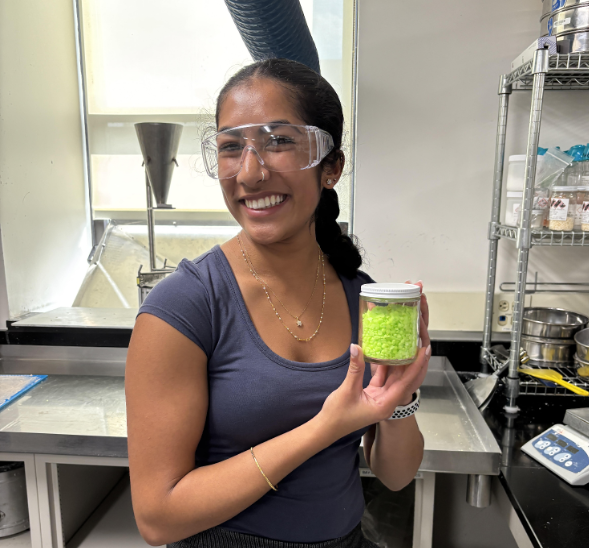 Smiling person in a lab wearing safety goggles, holding a jar of bright green granules.