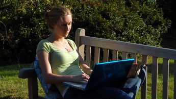 A student reclined on a bench working on a laptop