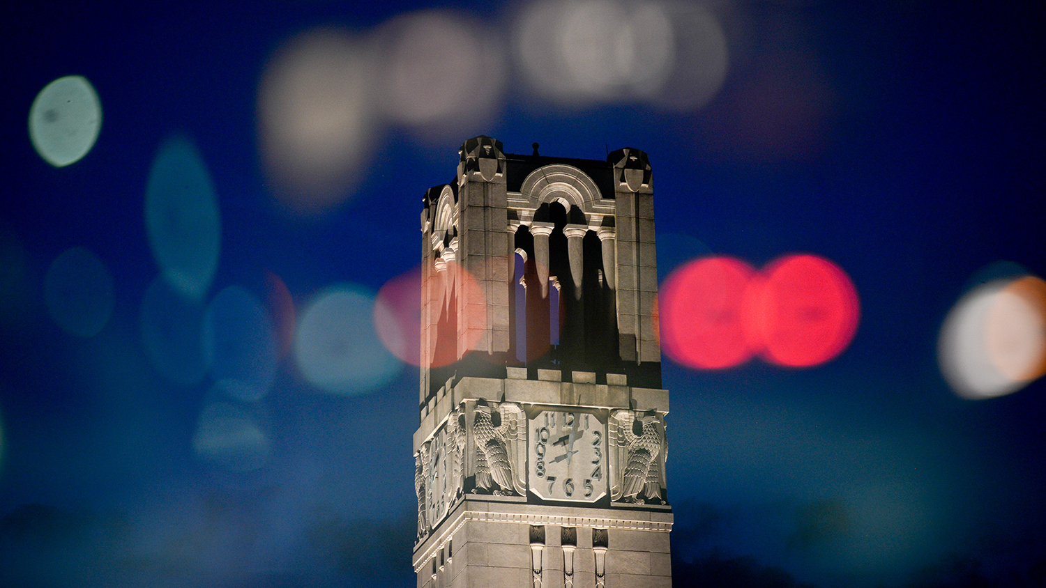 The NC State Belltower at dusk with colorful lights reflected around it