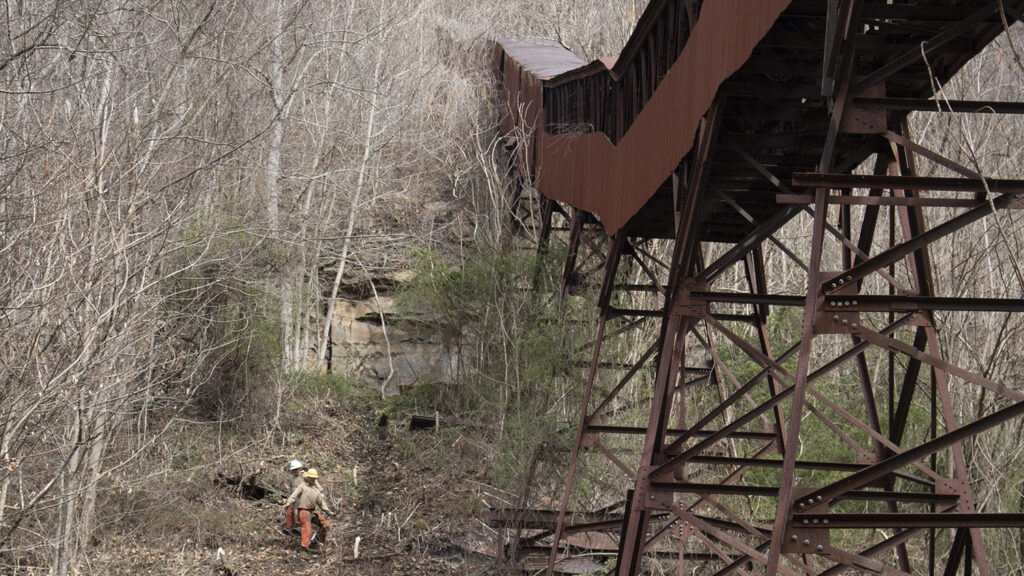 a large historic coal conveyor looms over workers clearing brush on a rocky, forested slope