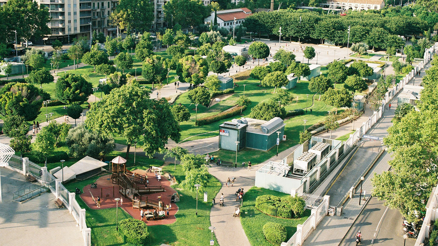 photograph is an aerial view of a city neighborhood, with busy streets running on either side of a park. The park has many trees, and there are large apartment and office buildings in the background.