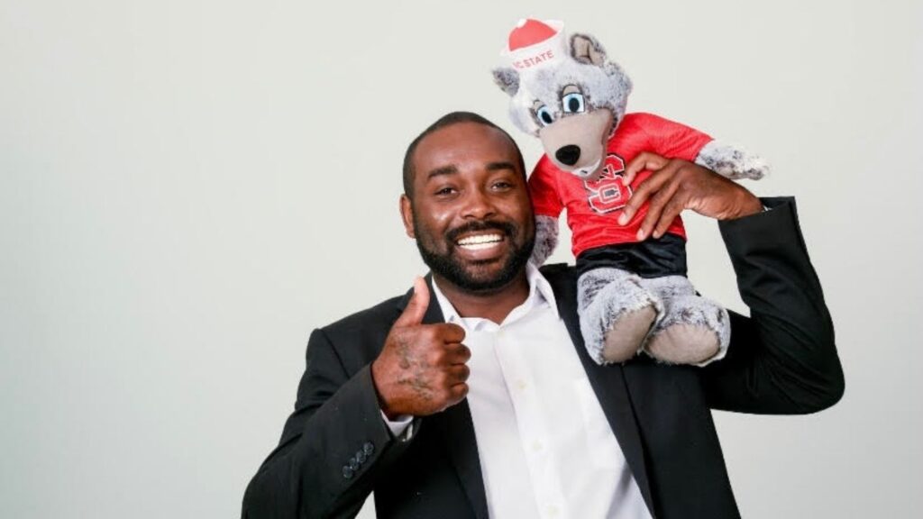 Rashawn King giving thumbs-up with NC State wolf stuffed animal on shoulder