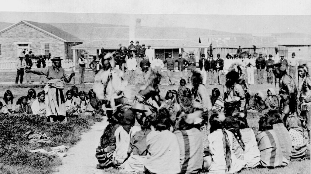 The Shoshone tribe at Fort Washakie, Wyoming in 1892. Some of the Shoshones are dancing as the soldiers look on. This is the last photograph of Chief Washakie, who is on the extreme left, standing and pointing.