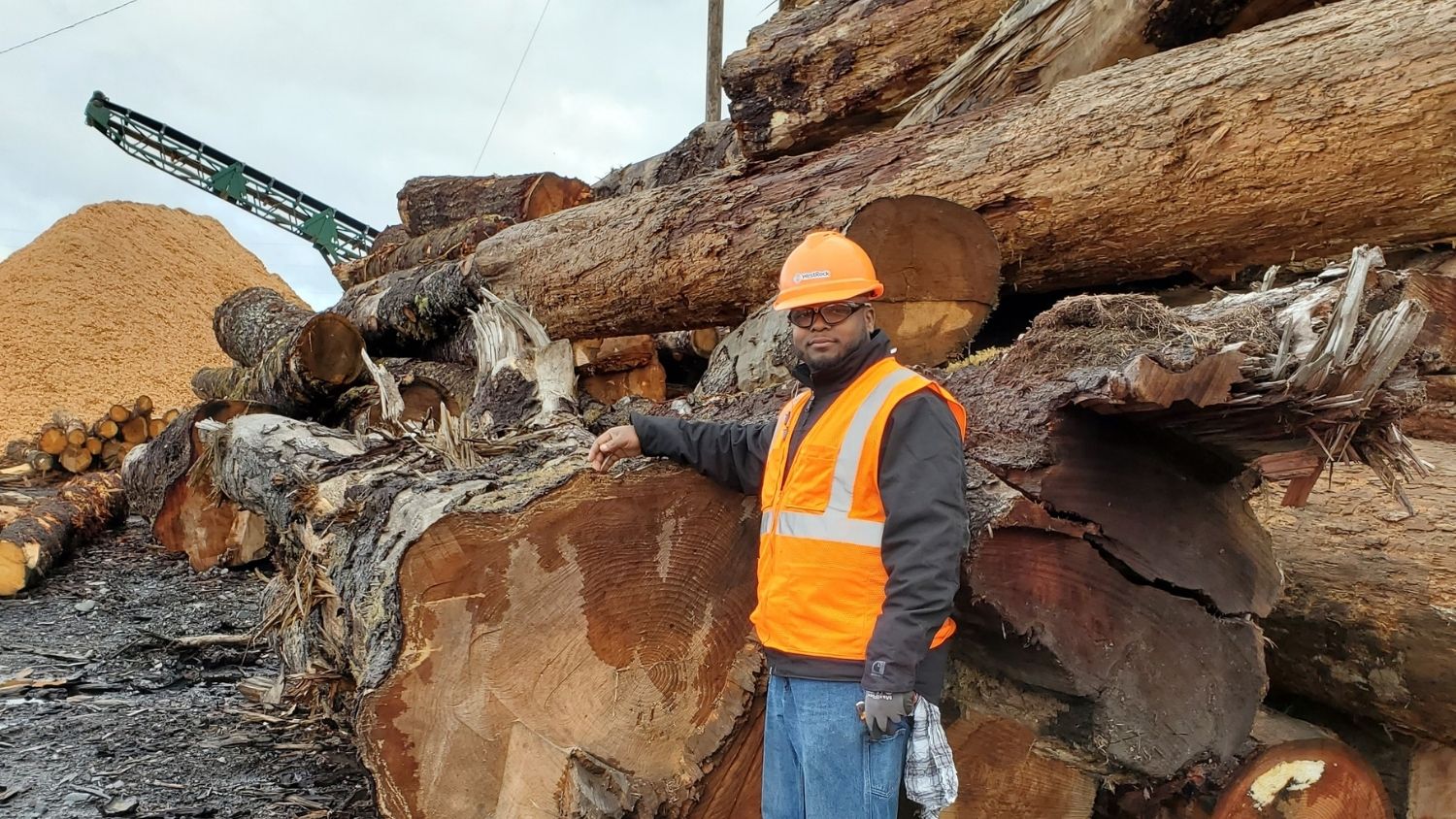 Elijah Gore stands in a hard hat in front of large logs
