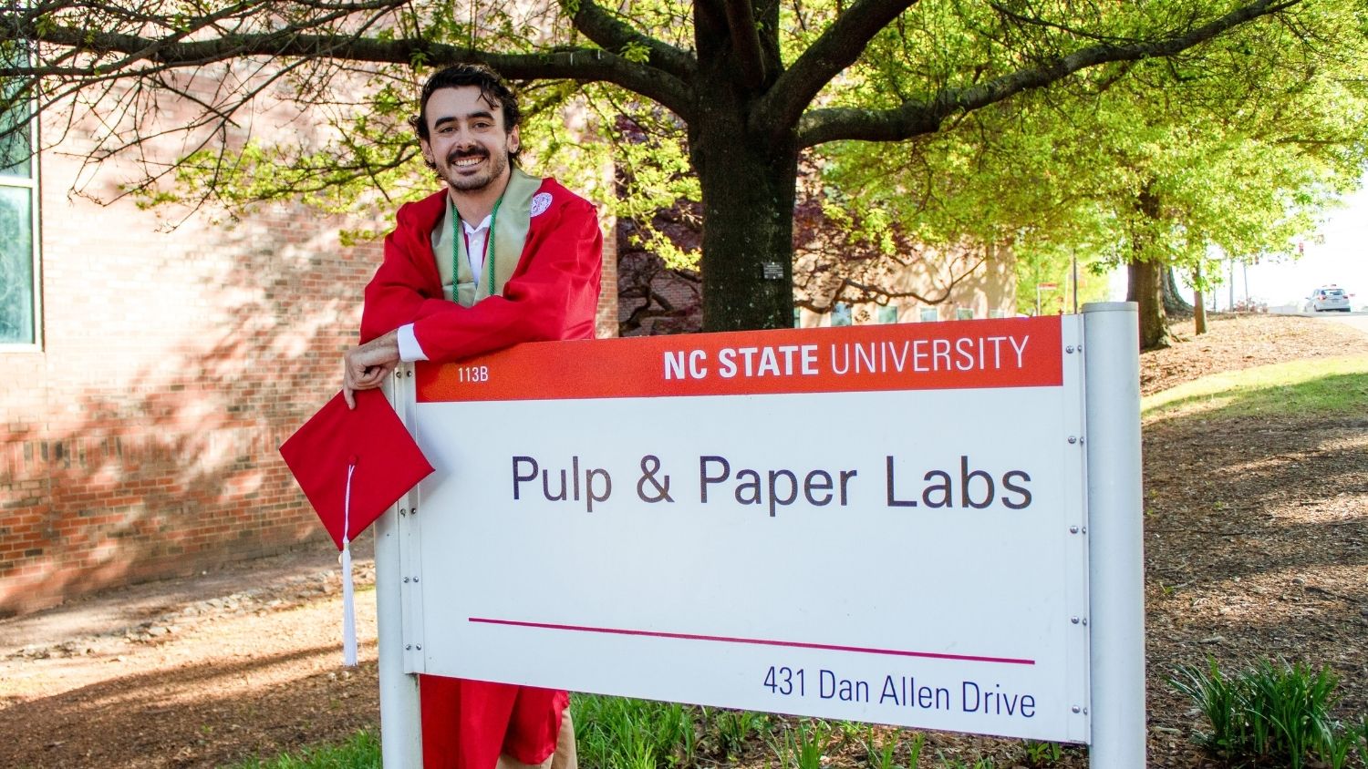 Brendan Tumpey stands in graduation robe by NCSU Pulp and Paper Labs sign