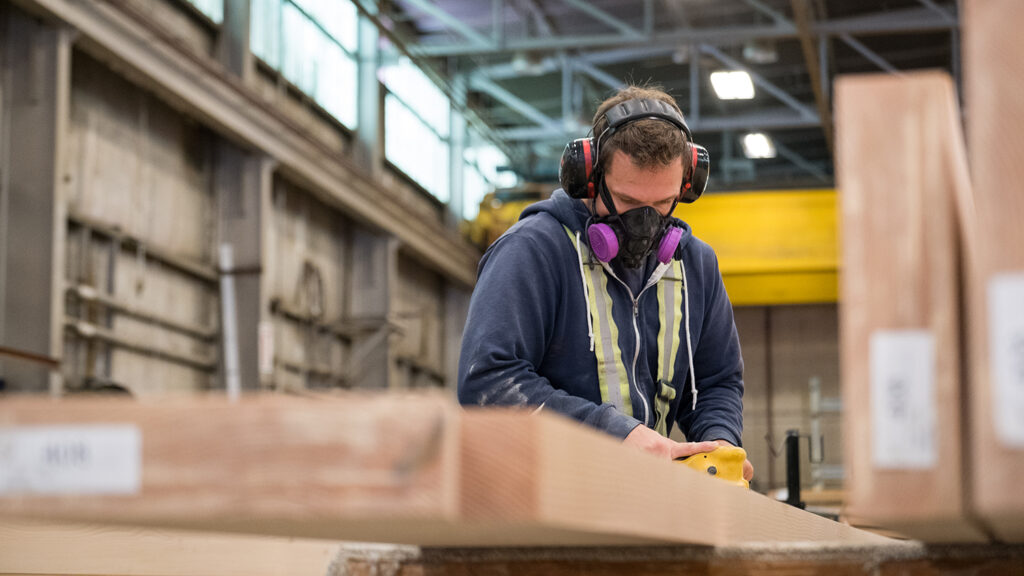 Man in a timber factory using an industrial orbital sander to smooth some wood. He has an industrial mask on protecting him.