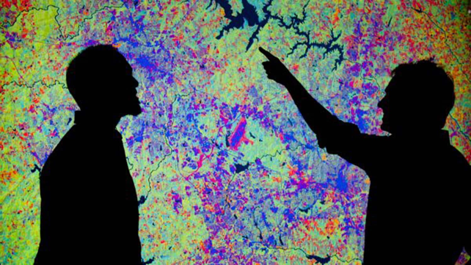 Two researchers appear as silhouettes in front of a screen showing a digital heat map filled with greens, blues, reds, oranges and various other shades of colors - Faculty Clusters Fuel a Culture of Excellence at NC State - College of Natural Resources at NC State University