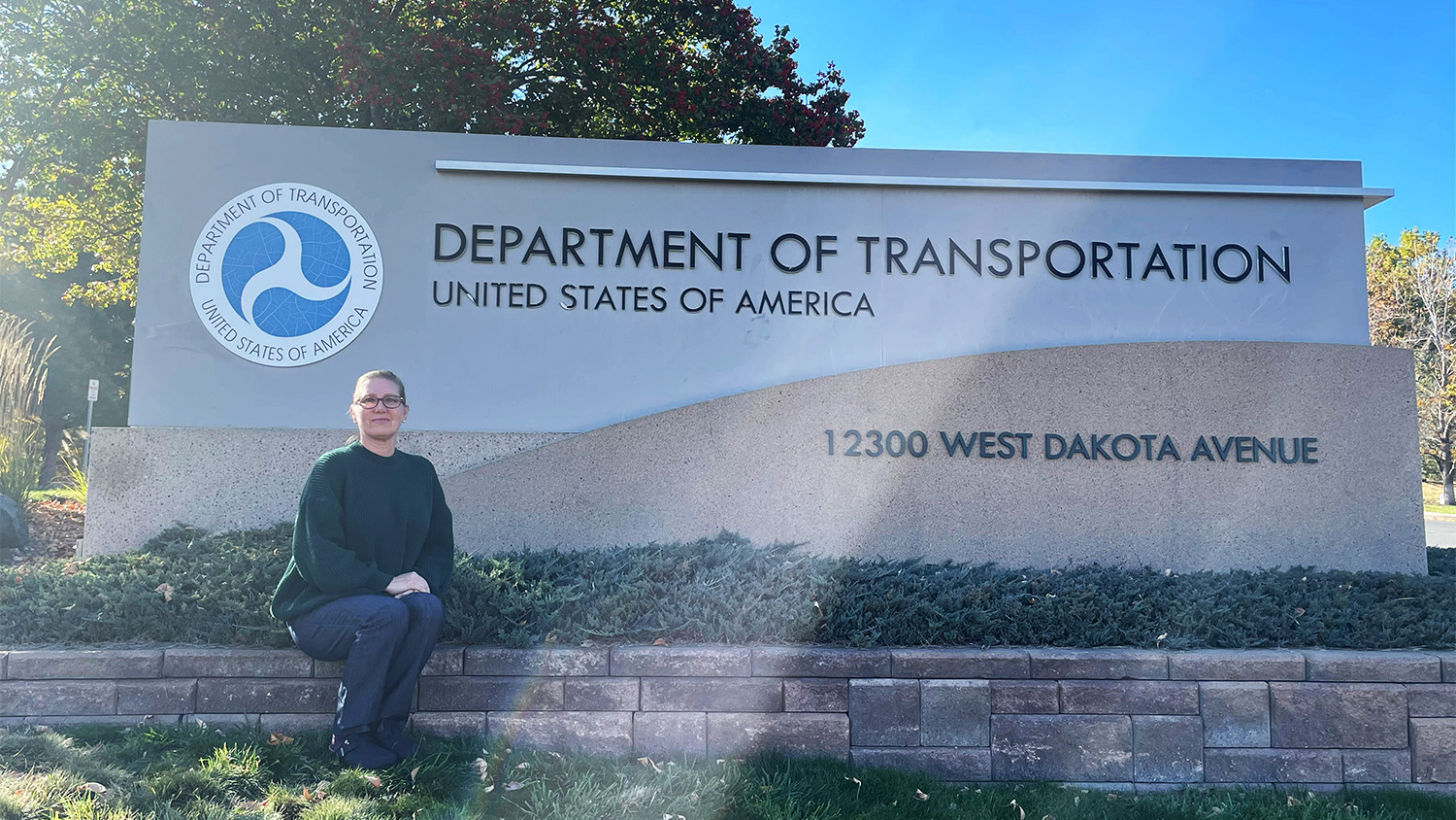 Chelly Sundermeyer sitting in front of Department of Transportation sign.