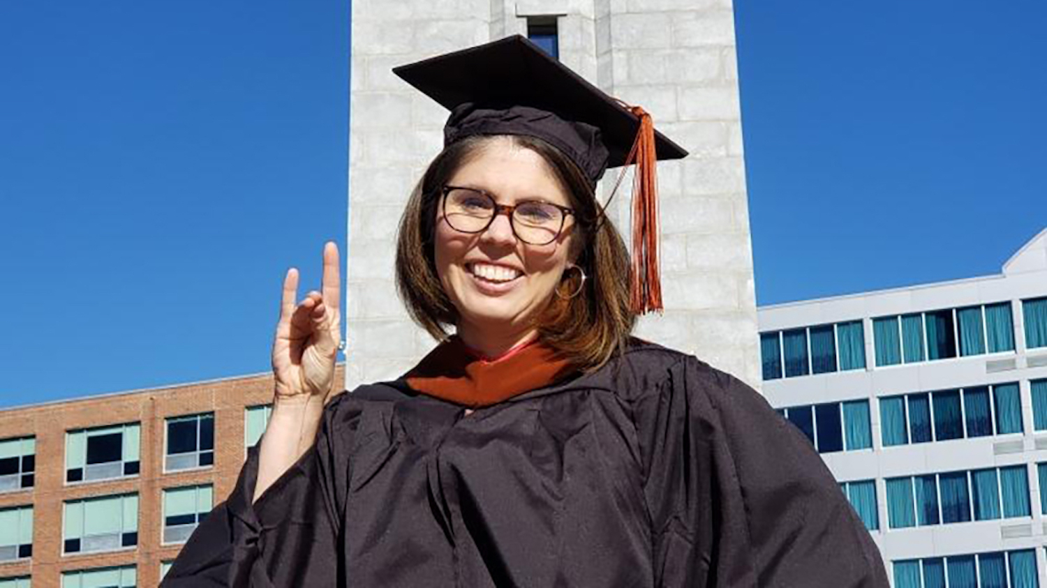 Kim Garrett gesturing with the“Wolfie” hand symbol in front of the Memorial Belltower on the NC State campus.