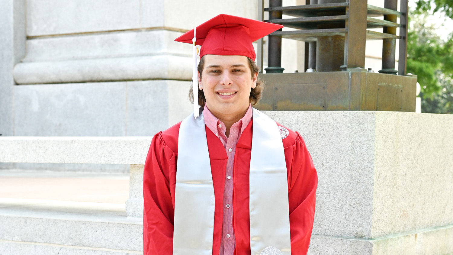 A student in his cap and gown stands in front of a concrete building.