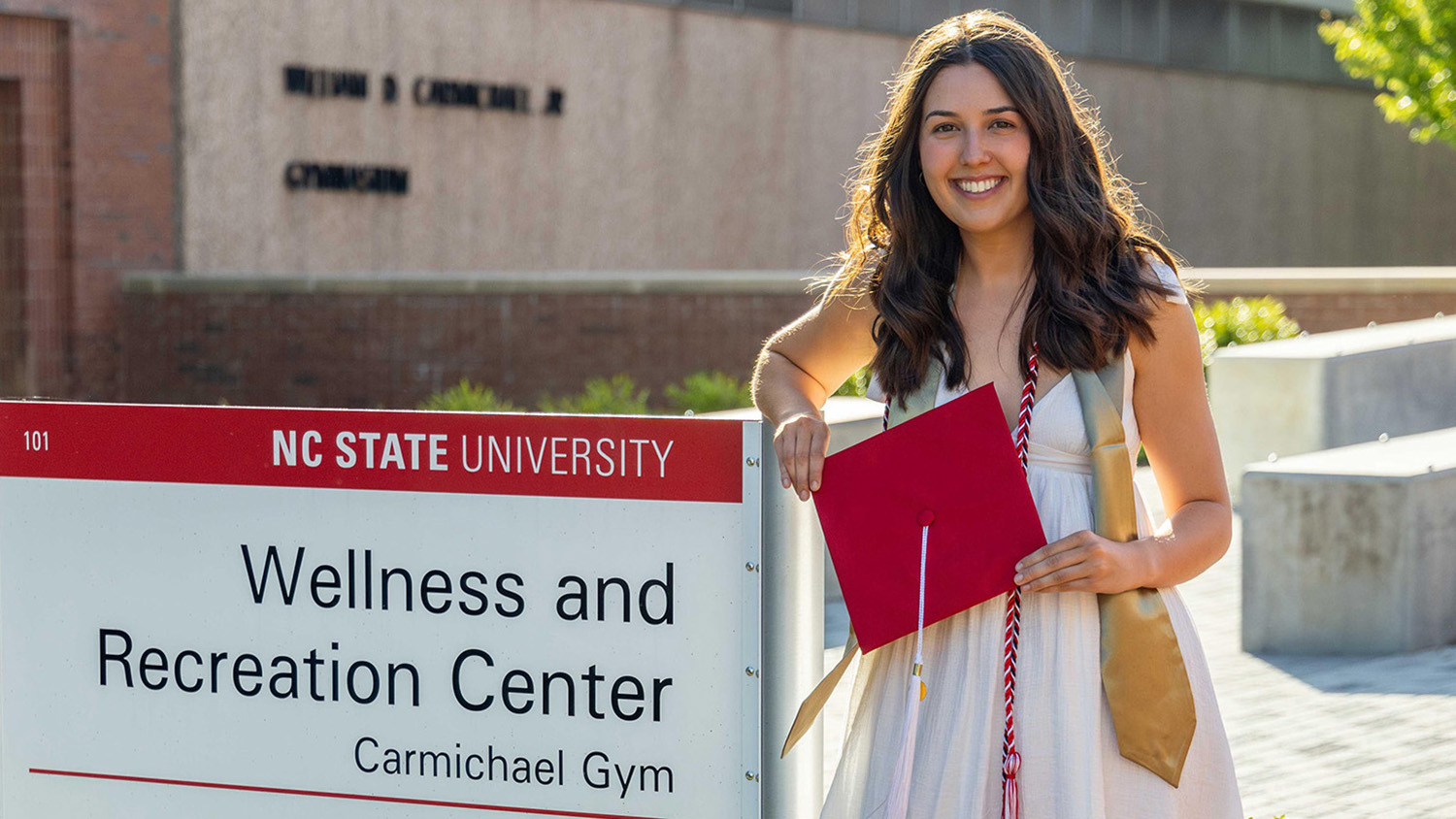 Julia Egler holding her garduation cap while posing beside the NC State Wellness and Recreation Center entrance sign.