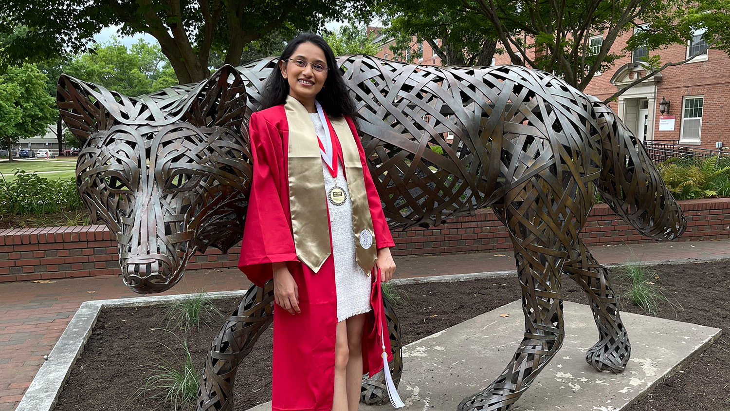 A student in her cap and gown stands in front of a bronze wolf statue.