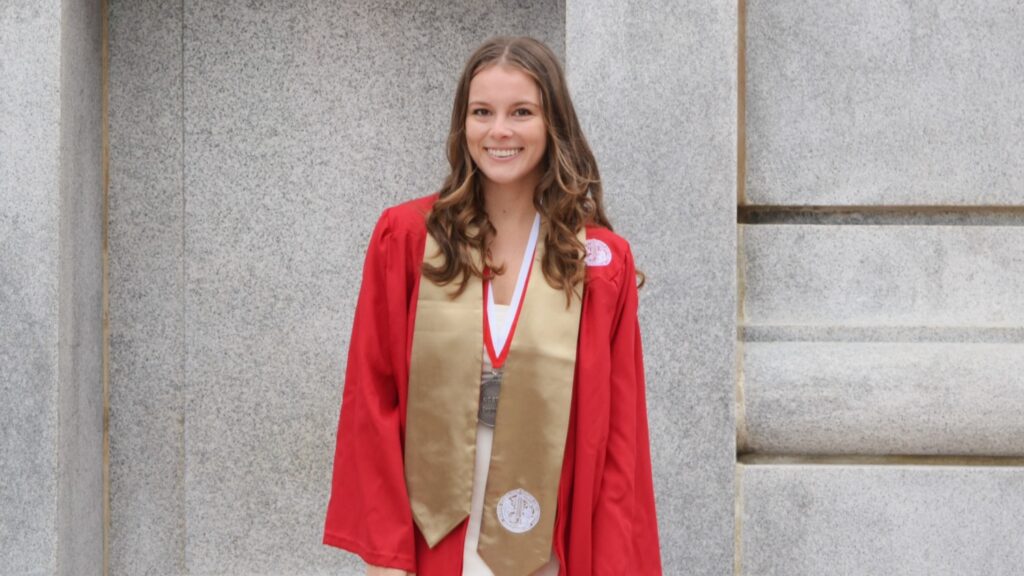 A woman in a red gown holding a red graduation cap in front a concrete structure.