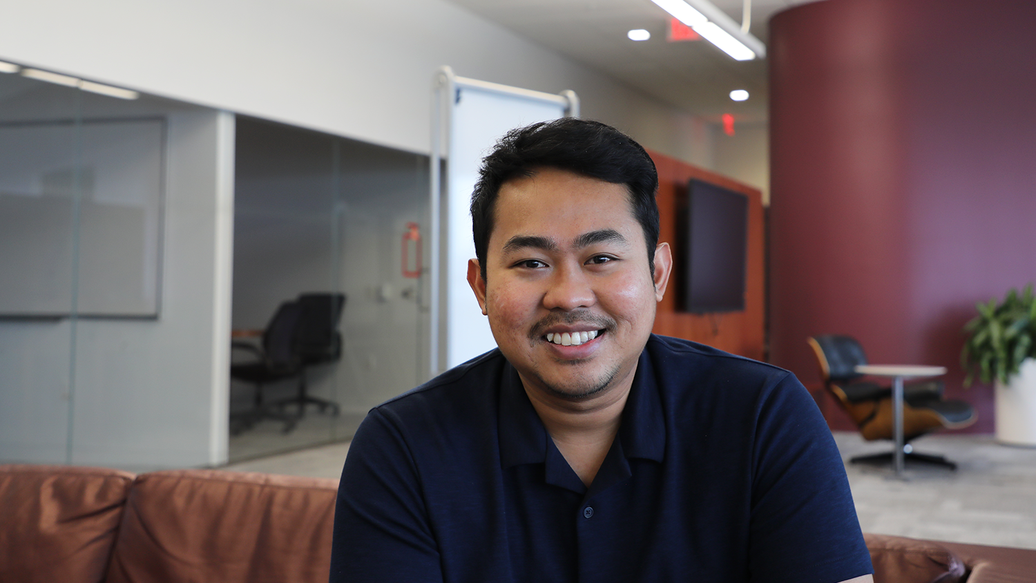 A man in a navy blue polo shirt sitting on a brown sofa in an office.