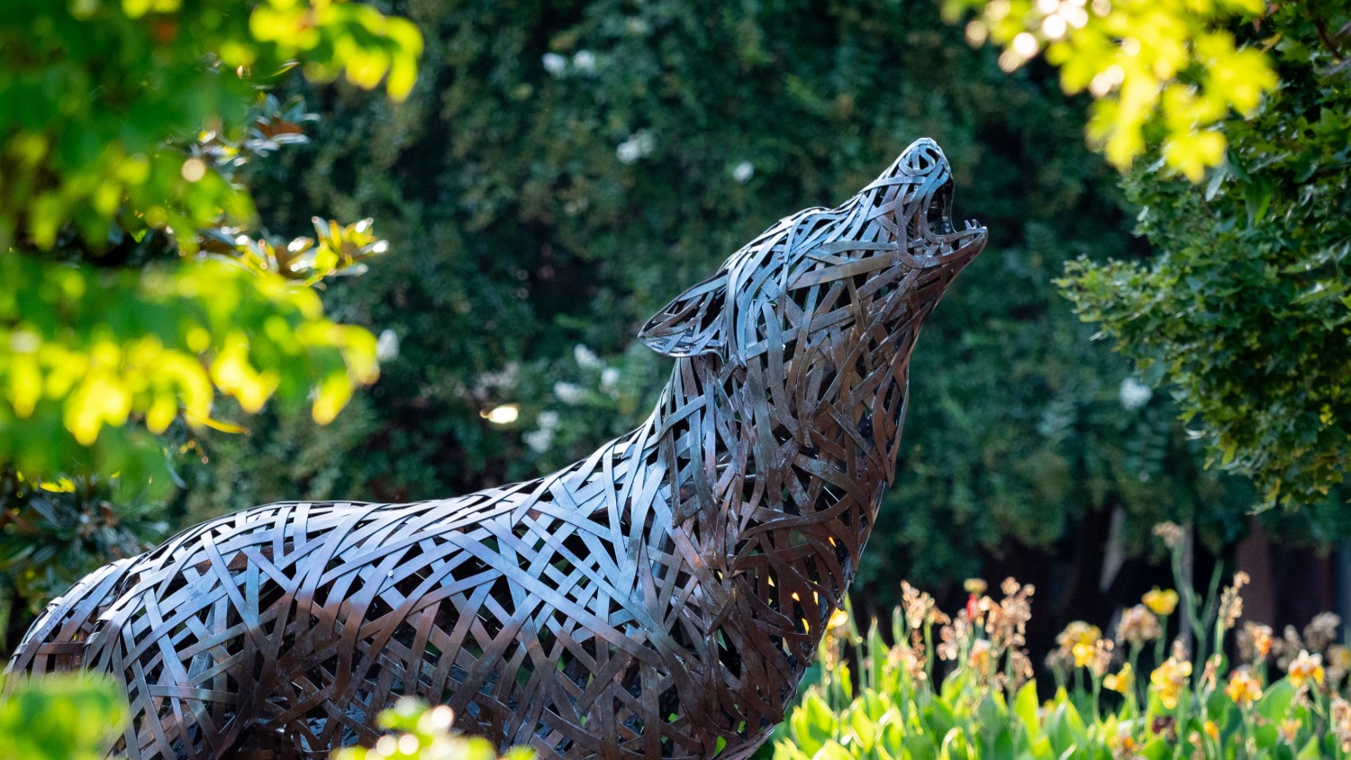 A copper statue of a wolf surrounded by trees and flowers.