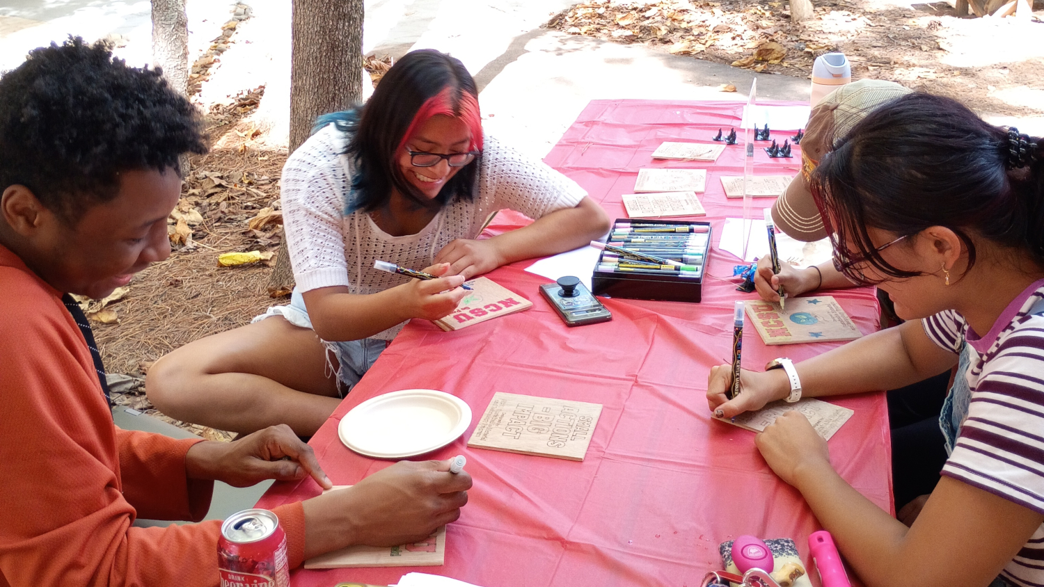 Students laugh and smile as they color wooden slabs with positive messages with markers on a picnic table. - Admissions - College of Natural Resources at NC State