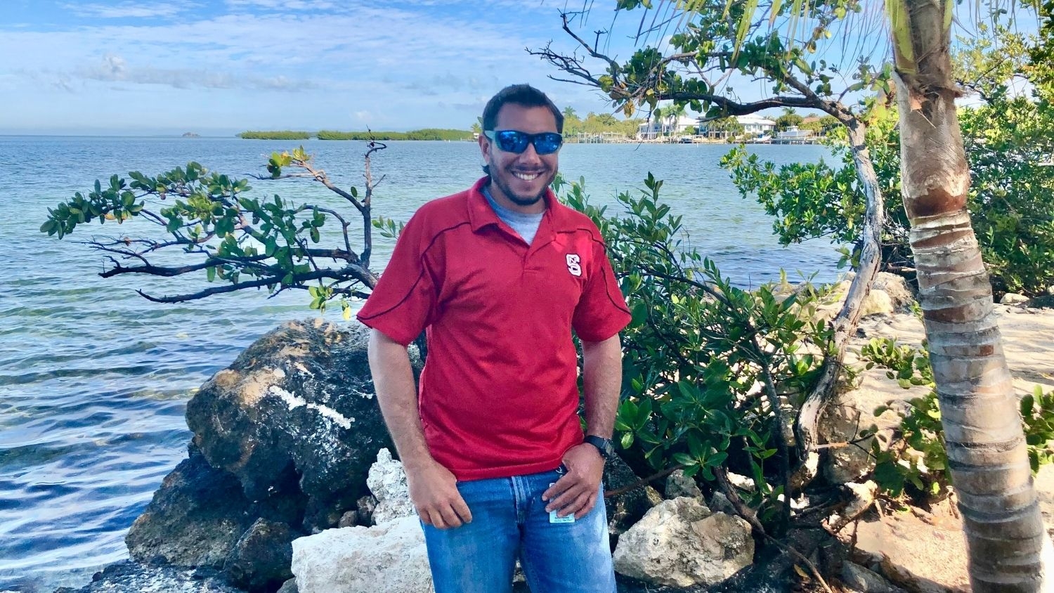 Daniel Parobok in NC State shirt in front of ocean and tropical tree
