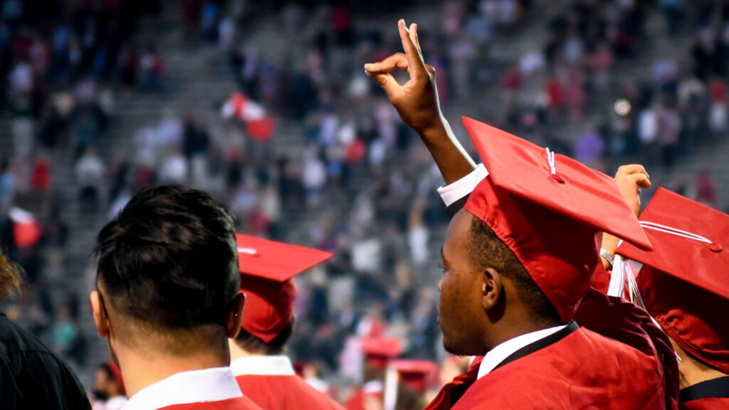 A graduate in a red cap and gown holds up the wolfie hand signal